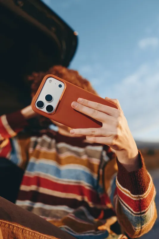 Woman holding smartphone for selfie, close up on hands, orange phone cover, striped sweater, outdoors with sky bokeh, lifestyle moment, casual travel mood