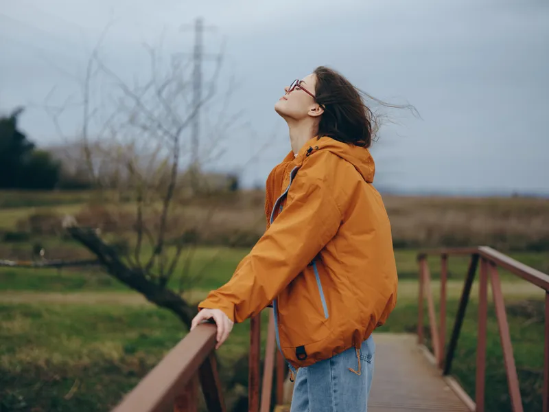 Woman lifestyle outdoors on wooden walkway, wearing orange jacket and jeans, hair lifted by wind as she looks up toward sky, calm nature with leafless trees and field in overcast light