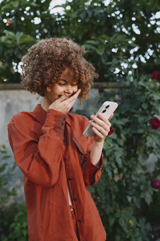 Casual woman in a rust jacket uses a smartphone outdoors in a lush garden, capturing a candid lifestyle moment with warm natural light and a thoughtful expression, against a leafy backdrop