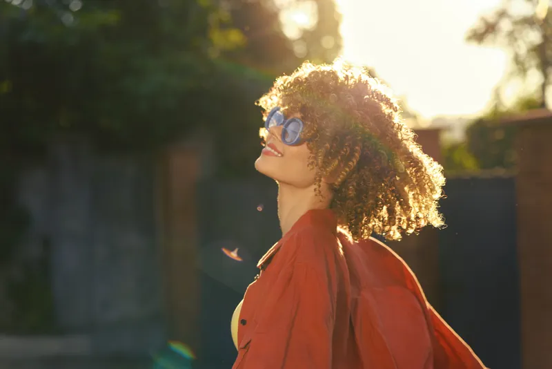 Cheerful woman outdoors, sunlit moment, wearing stylish sunglasses, curly hair flowing, warm breeze, casual outfit, joyful expression, vibrant colors, natural background, lifestyle portrait