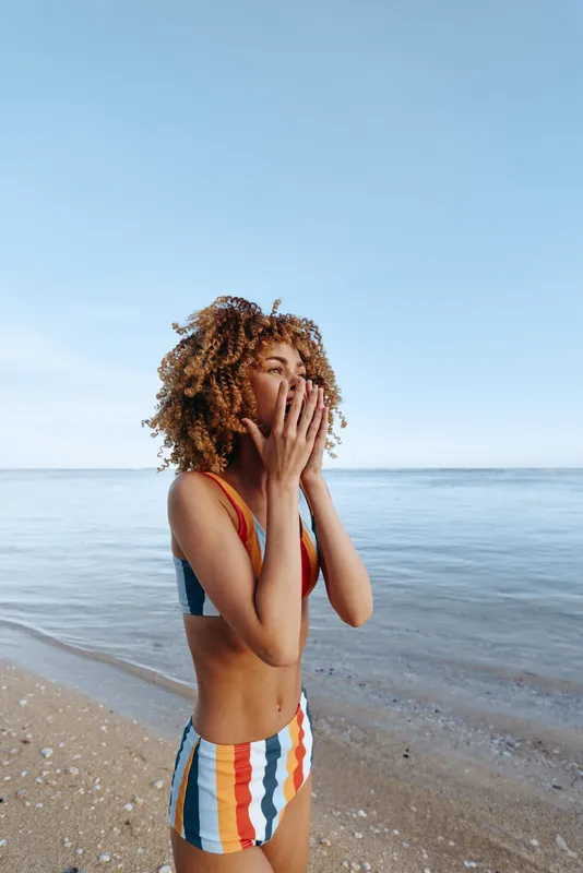 Woman beach ocean curly hair covering mouth hands on sandy shore, striped swimsuit, windy breeze, calm waves at horizon, bright summer sky, quiet surprise, copy space