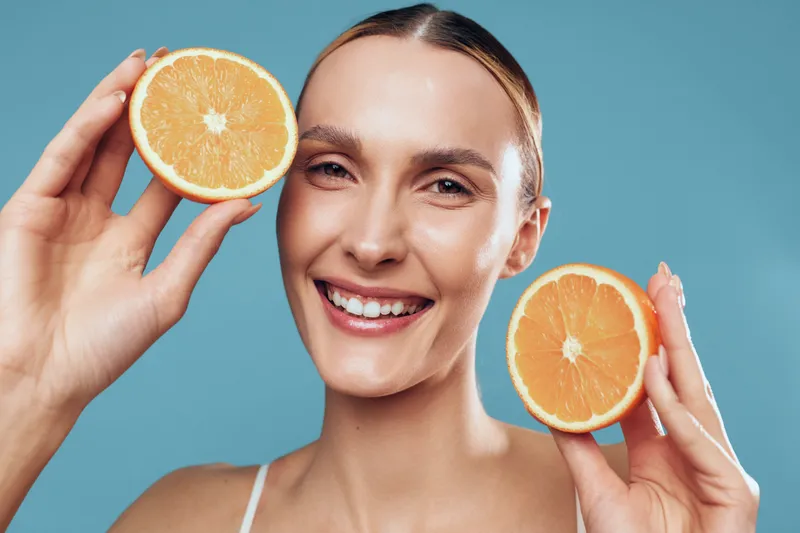 A smiling woman holds two orange halves near her face against a blue backdrop, conveying freshness, citrus energy, and healthy living through vibrant color and clear lighting.