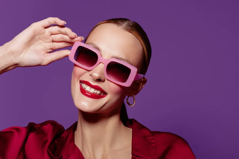 Smiling woman with pink sunglasses and red lipstick poses against purple background. Fashion portrait with bright colors, trendy style, and confident mood in studio lighting.