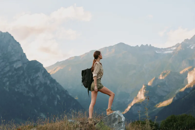Young woman with backpack standing on rock in mountain landscape. Outdoor adventure, hiking, and lifestyle concept. Natural light highlights scenic view and active travel.