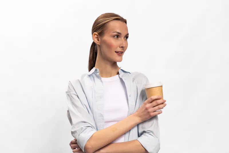 Young woman with braided hair holds a paper cup, wearing casual white shirt and light jacket. She looks to the side with a thoughtful expression on white background.