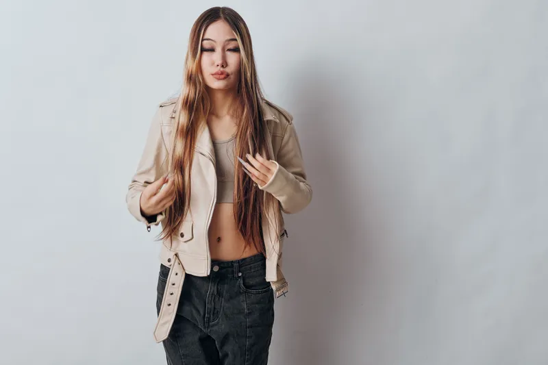 Young woman with long hair wearing beige jacket and crop top standing against white background. Casual style, fashion outfit and confident relaxed pose indoors for portrait.