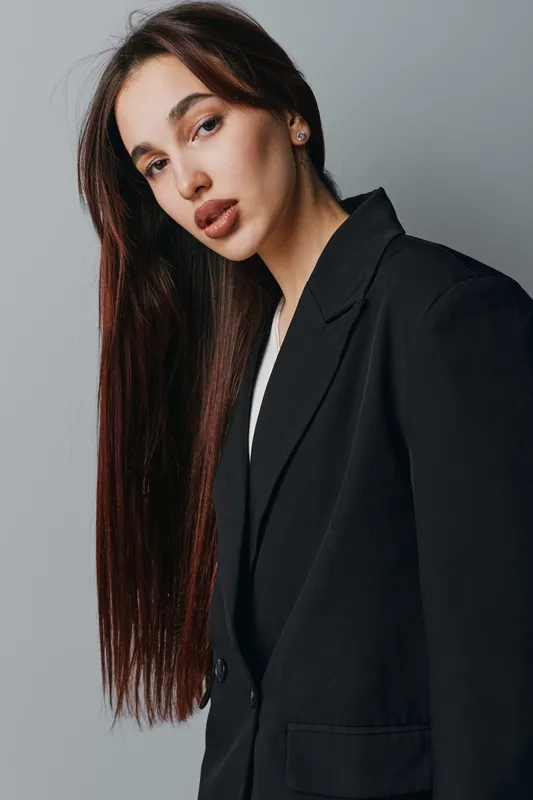 Woman with long brown hair and natural makeup wearing black blazer posing against gray background. Portrait of confident female model with straight hair and neutral expression in studio lighting.