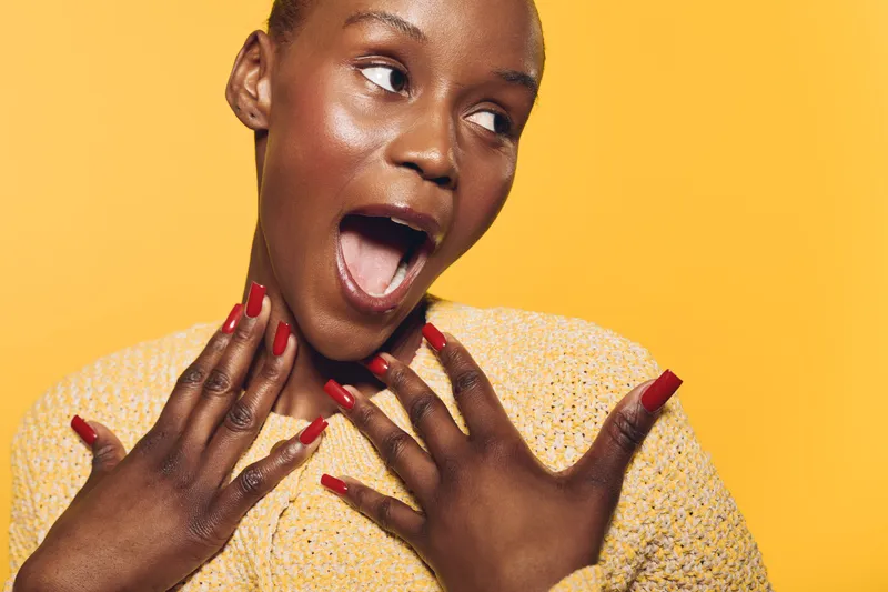 Surprised young black woman with dark skin and red nail polish looks amazed with open mouth. Bright yellow background and casual sweater create lively, expressive portrait concept.