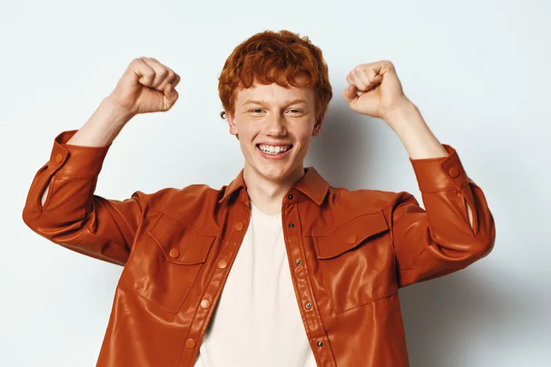 Young man with red hair smiling and raising fists in a celebratory gesture wearing brown shirt on white background. Happy expression and confident body language.