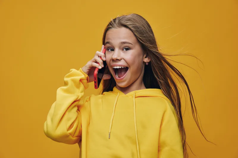 Girl with long hair wearing yellow hoodie talking on red smartphone and smiling with open mouth, isolated on yellow background with happy and excited expression.