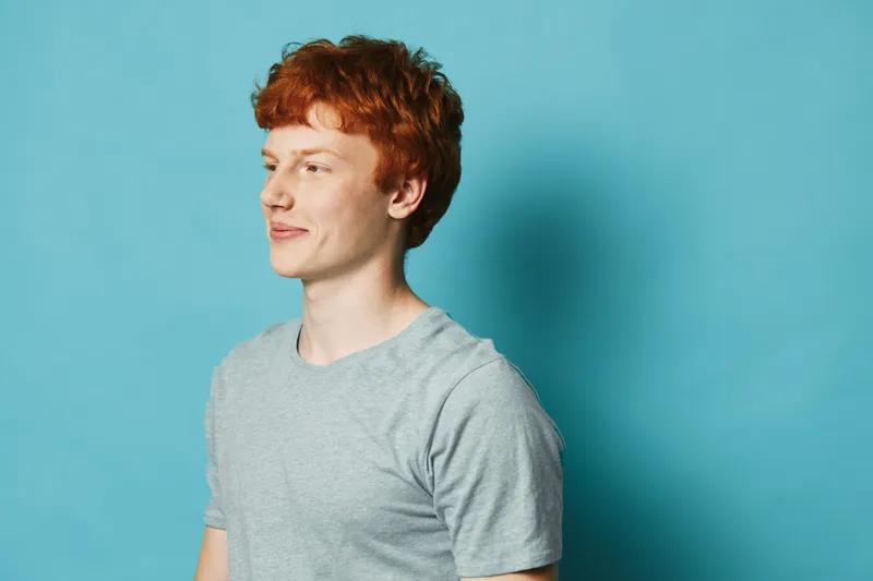 Young man with red hair and freckles wearing a gray t-shirt standing against blue background looking to side with calm expression and relaxed posture in studio setting.