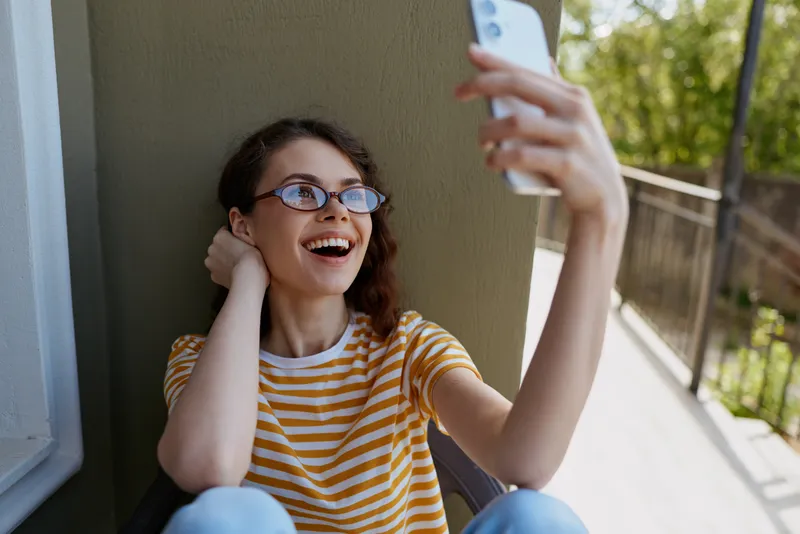 Bright, smiling woman takes a selfie on a sunny balcony, wearing a striped tee and glasses. Casual lifestyle moment captured for social media and home life.