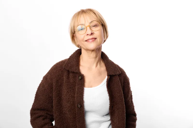 Senior woman with glasses smiling in a studio portrait, white background, brown coat and white sweater, relaxed face and calm expression, soft light, friendly mood