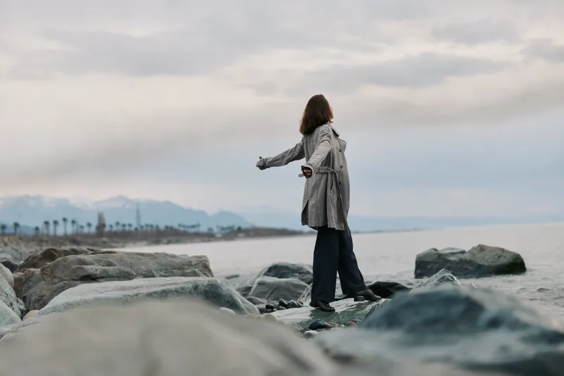 A woman in a long coat stands on rocks by the sea with arms outstretched, looking at the cloudy sky over distant mountains. Concept of freedom, nature, and solitude outdoors.