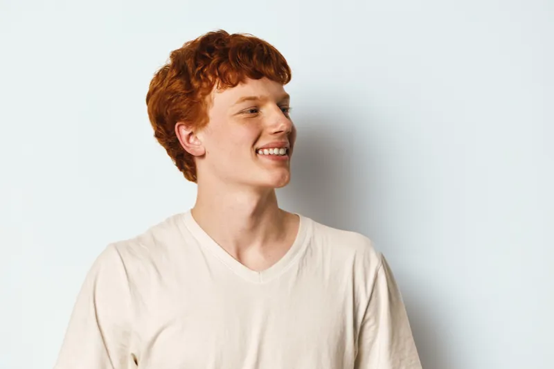 Young man with red hair smiling and looking sideways wearing light t shirt isolated on white background. Portrait of cheerful male with casual style and happy expression.