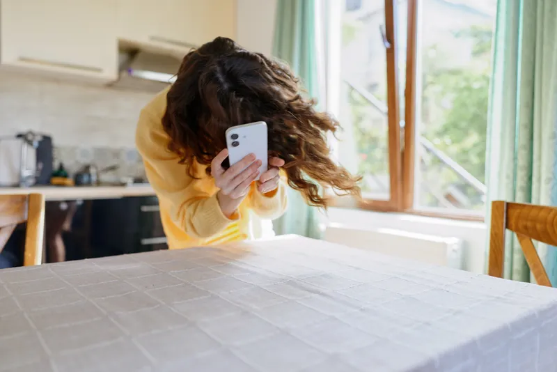 Young woman with wavy hair in a yellow sweater using a smartphone at a kitchen table. Bright natural light creates a cozy home scene, casual lifestyle, and a candid moment.