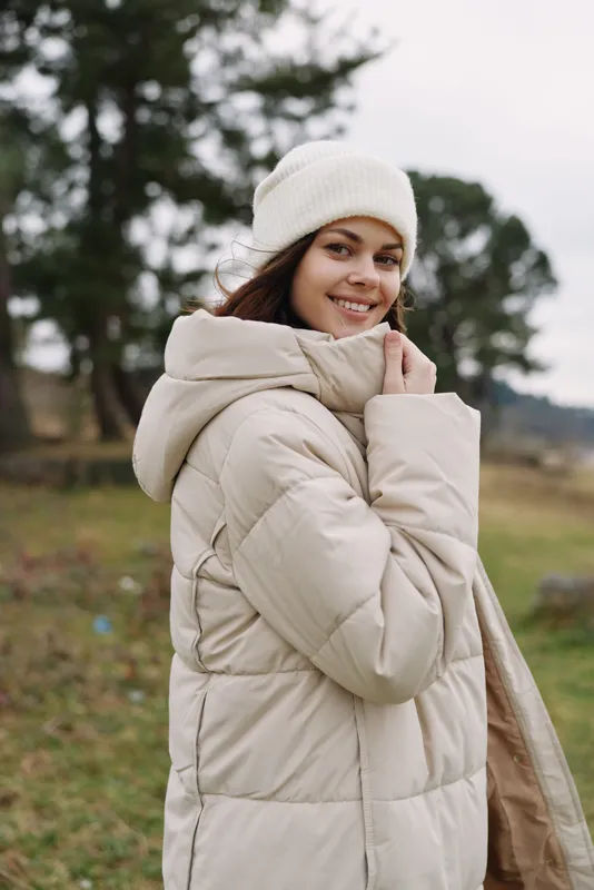 Winter outdoor portrait of a smiling woman in a beige coat and knit hat, posing in a field with trees, exuding cozy fashion and seasonal calm today
