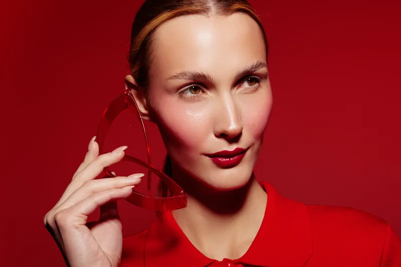 Portrait of a stylish woman with red lipstick and red outfit holding red sunglasses on red background. Concept of fashion, beauty, elegance, and modern style with vibrant colors.