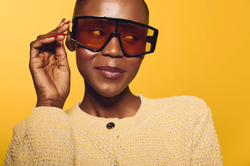 Portrait of a confident black woman with dark skin wearing oversized sunglasses and a yellow sweater against a bright yellow background. Concept of style, fashion, and self-expression.