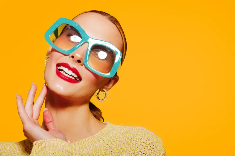 Smiling woman with large blue sunglasses and red lipstick poses against bright yellow background. Fashion portrait with vibrant colors and joyful expression. Concept of style and beauty.