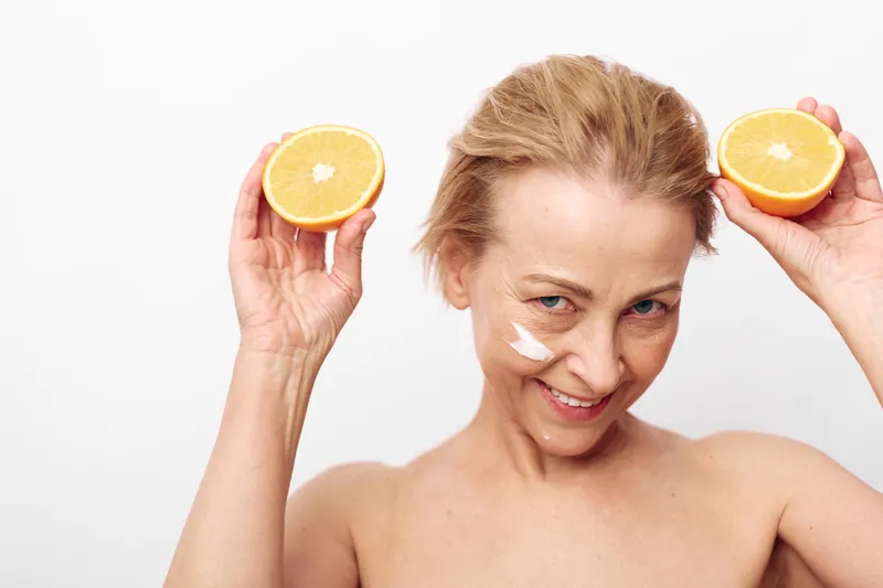 Smiling mature woman with cream on cheek holds two orange halves near her face. Bright white background highlights natural skincare, healthy lifestyle, and vitamin c beauty concept.