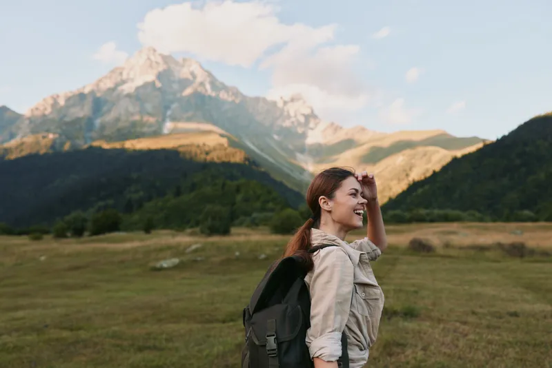 A young woman with a backpack wanders through a green meadow with towering mountains behind, enjoying a peaceful outdoor adventure in clear weather.