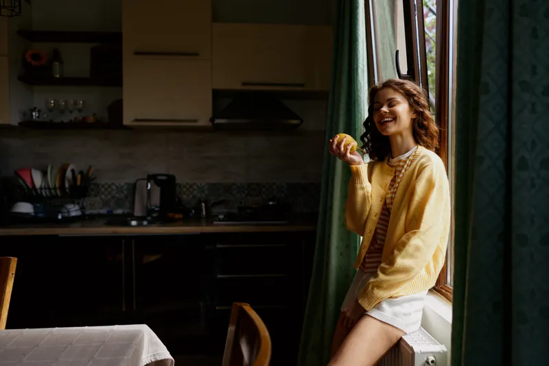 A cheerful woman in a yellow sweater sits by a sunlit kitchen window, enjoying a moment of casual relaxation. Bright interior, home comfort, and everyday warmth convey a relatable lifestyle moment.