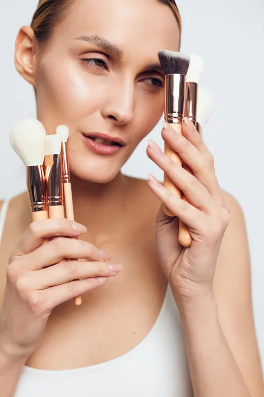 Young woman with blonde hair holds makeup brushes close to her face while applying powder with a focused expression against a plain white background.