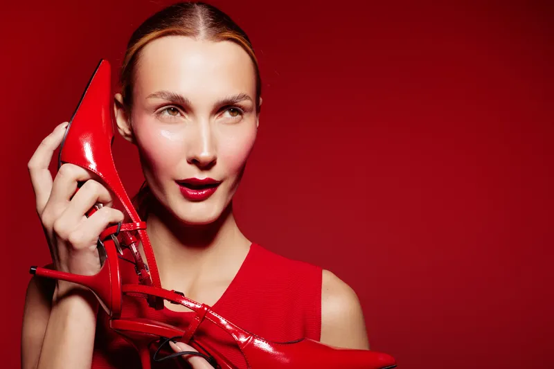 Woman in red dress holds red high heel shoes near her face against red background. Fashion concept with bright colors and stylish accessories in studio lighting.