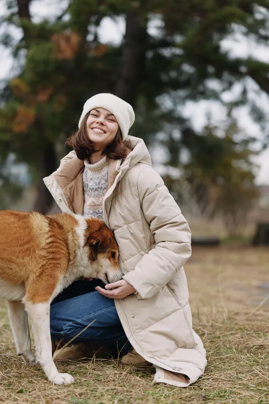 Woman in a light beige coat kneels with a friendly dog in a park, smiling under a knit hat as autumn leaves fall around them, capturing warm outdoor companionship