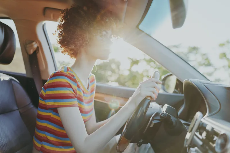 Young woman with curly hair drives a car on a sunny day, wearing a striped shirt, enjoying a carefree moment behind the wheel.