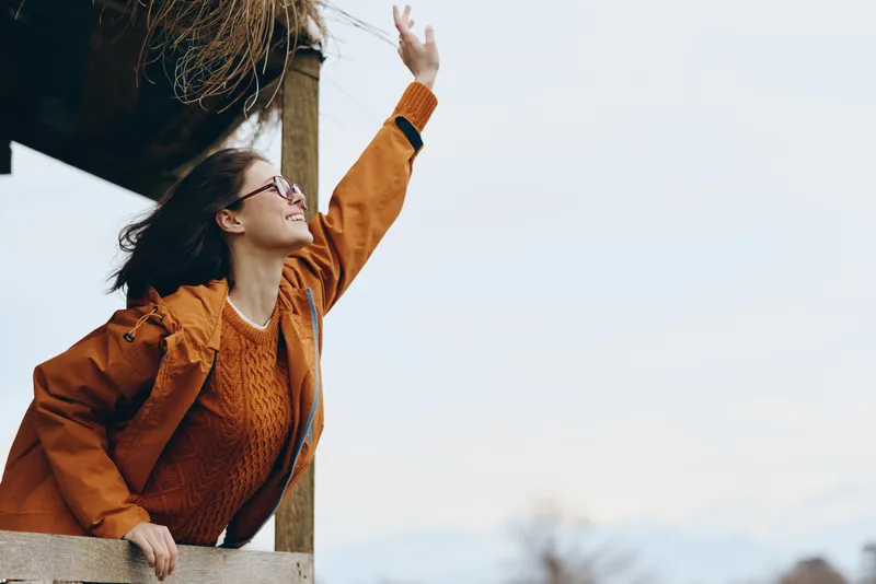 Woman lifestyle portrait outdoors in autumn coat and glasses, smiling and resting by a post, one arm raised, soft daylight, calm moment, blurred background