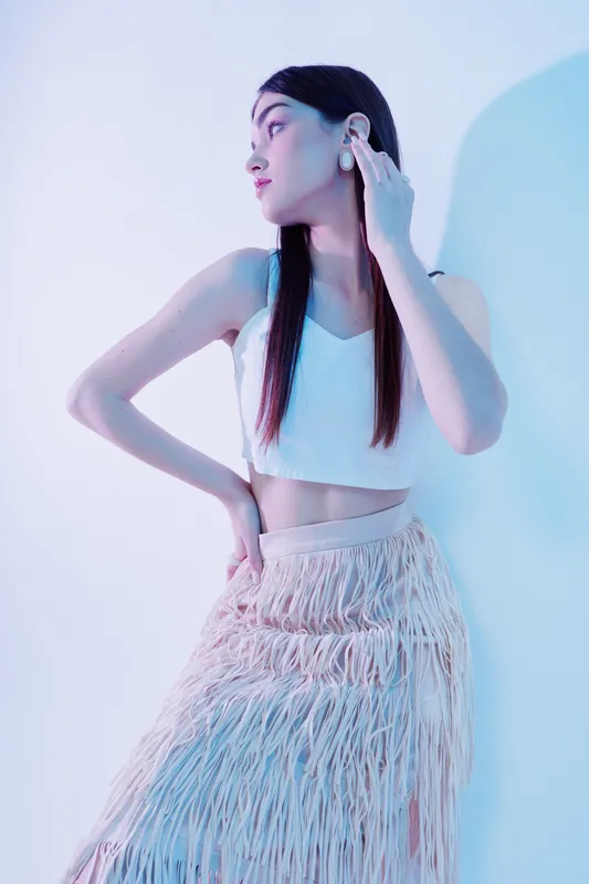 Young woman in a white crop top and fringed skirt poses with hand on hip and looks sideways. Studio shot with soft blue and pink lighting. Concept of beauty, fashion, and style.
