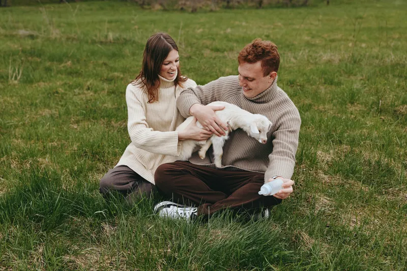 Goat kid and couple share a warm moment on grassy outdoors, a candid scene of family lifestyle, cuddle, love, and connection in a peaceful nature setting with soft light.
