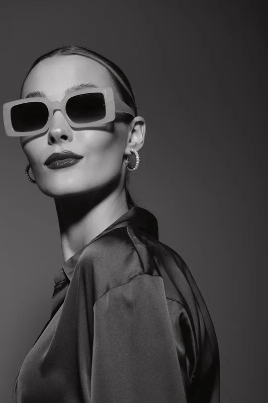 Stylish white woman with pale skin wearing oversized sunglasses and hoop earrings poses confidently in silky blouse. Black and white studio portrait with fashion and elegance concept.