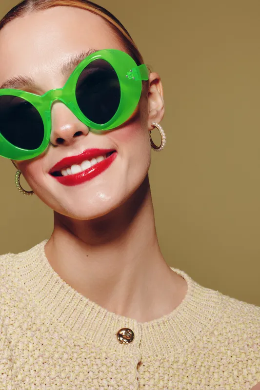 Smiling woman with bright red lipstick wearing large green sunglasses and hoop earrings. She wears a beige knitted sweater against a plain olive background. Fashion and style concept.