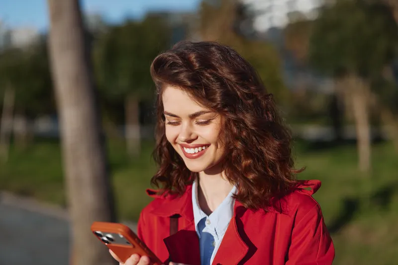 One smiling brunette woman in a red coat looks at her phone in a sunlit park with green trees in the background, a soft breeze and relaxed posture.