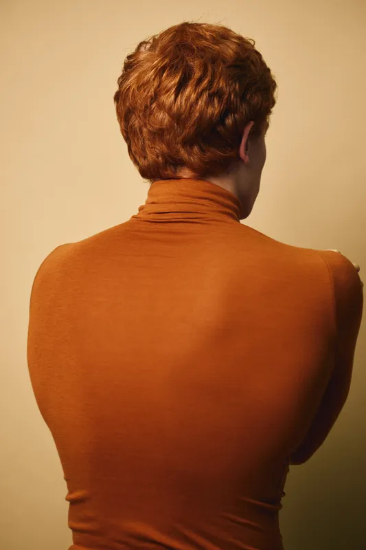Young man with curly red hair in brown turtleneck sweater seen from back with soft warm lighting on beige background. Casual style and minimalistic portrait composition.