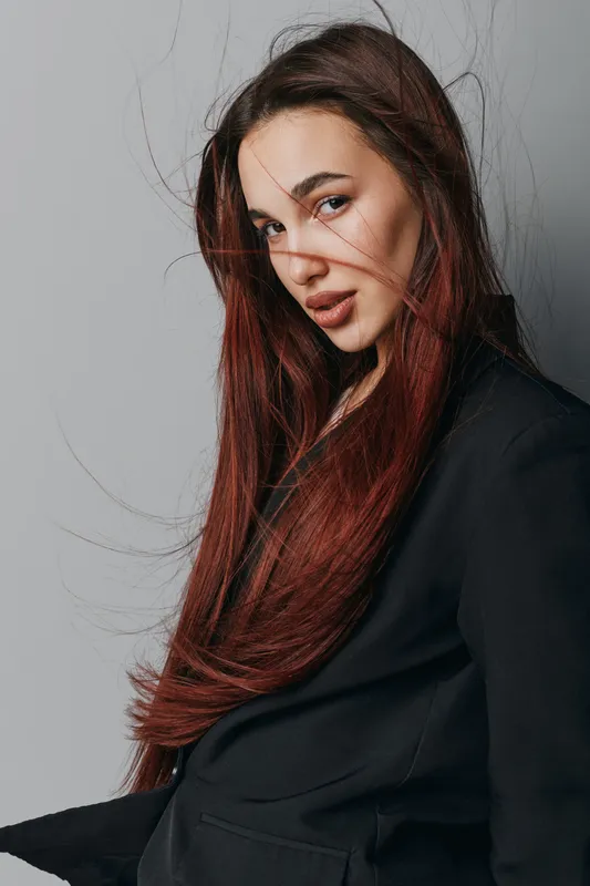 Portrait of young woman with long red hair wearing black jacket posing against gray background. Attractive female model with natural makeup and confident expression in studio lighting.
