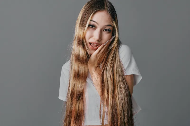 Young woman with long straight hair posing with hand on face in white t shirt against neutral background, beauty and fashion portrait with natural makeup and casual style.