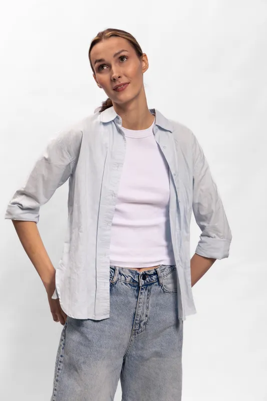 Young woman in casual outfit with light shirt and jeans stands relaxed on white background. Concept of casual fashion, youth, confidence, and simplicity with natural expression.