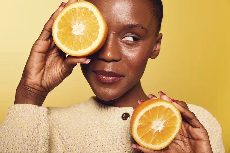 Close-up of a smiling dark skinned African woman holding two orange halves near her face. Concept of healthy lifestyle, natural beauty, fresh fruit, and wellness on yellow background.