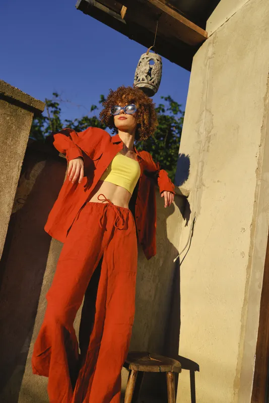 woman lifestyle outdoor fashion red yellow sunglasses pose wall rustic, cheerful woman outdoors in sunlit moment, bold color palette, relaxed pose and urban charm