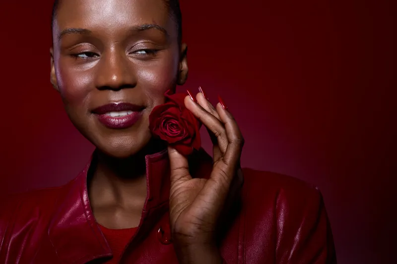 Close-up of a smiling black woman with dark skin holding a red rose near her face. She wears a red leather jacket with a red background. Concept of beauty, elegance, and romance.