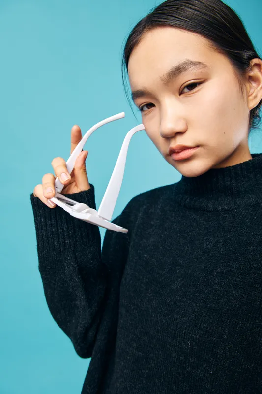 Young woman wearing black sweater holding white sunglasses near face posing against blue background, casual fashion look with serious expression and minimalistic style for portrait.