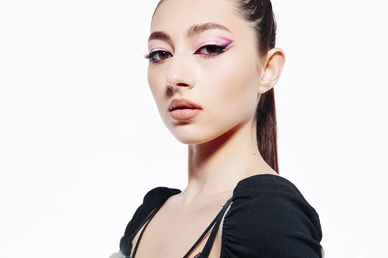 Young woman with bold pink eyeliner and glossy lips looks confidently at camera. Clean white background for beauty fashion concept with creative minimalist makeup style.