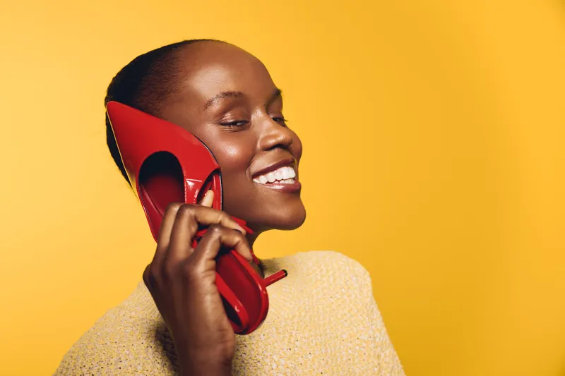 Smiling black woman with dark skin holds red high heel shoe to ear like phone, wearing beige sweater on yellow background. Concept of creativity, fashion, and playful communication.
