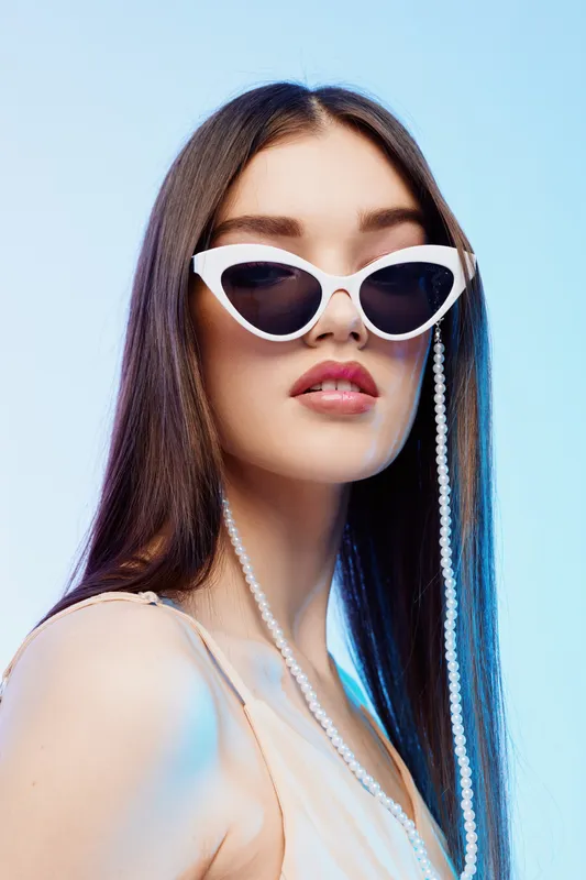 Young woman with long dark hair wearing white sunglasses and a pearl necklace poses against a blue background. Concept of beauty fashion and modern style.