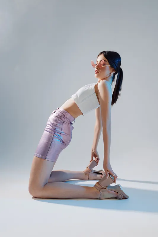 Young woman in a white crop top and shiny pink skirt poses on her knees in studio light. Fashion concept focused on beauty, style, and modern trends with soft shadows.