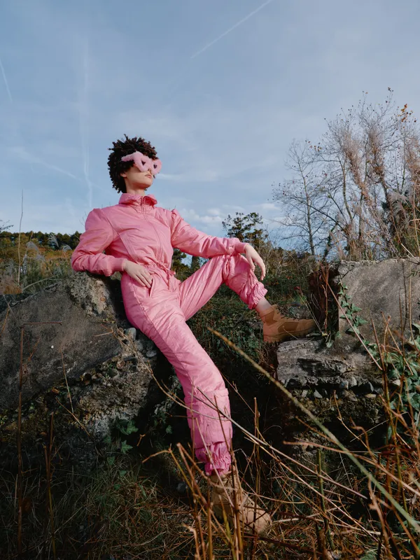 Person in a pink suit posing confidently outdoors on rocky terrain, highlighting fashion, adventure, and bold style amid a natural landscape under a clear blue sky